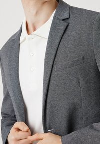 Blazer grigio tessuto sopra una polo bianca, con colletto, tre bottoni e un sottile dettaglio sulla tasca. Il tessuto appare morbido.