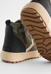 Baskets montantes texturées vert foncé et noires avec une semelle en caoutchouc beige clair et un motif de semelle en zigzag ; elles présentent une fermeture éclair sur le côté pour un accès facile.