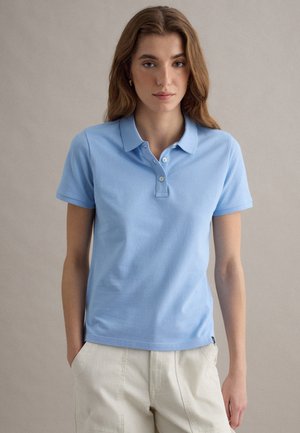 Jonge vrouw met lichtbruin haar draagt een lichtblauw poloshirt met korte mouwen en beige broek, staand tegen een effen achtergrond.