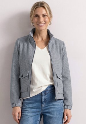 Uśmiechnięta kobieta w szarej, kratowanej kurtce na suwak, na białej koszuli i niebieskich jeansach, stojąca na jasnym, jednolitym tle.