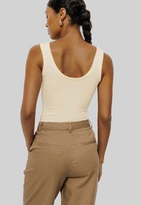 Débardeur beige avec des bords festonnés, porté avec un pantalon taille haute marron doté de poches arrière, sur fond gris.
