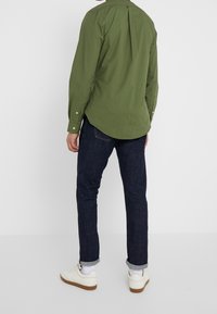 Olivově zelená košile s dlouhým rukávem a záhybem na zádech, doplněná tmavě modrými džínami a bílými teniskami, s ohrnutými manžetami.