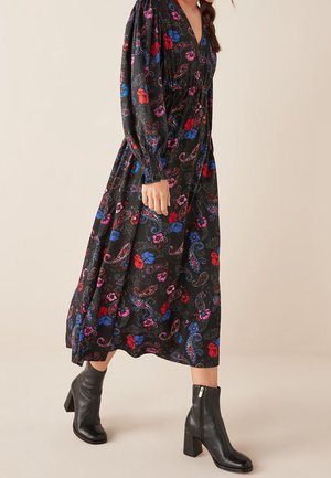 Frau trägt ein schwarzes Midikleid mit Blumenmuster in Rot, Blau und Pink, kombiniert mit schwarzen Stiefeletten mit Absatz, vor neutralem Hintergrund.