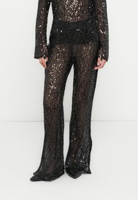 Pantalon noir à sequins avec une taille ajustée, des jambes évasées, une superposition transparente et une surface texturée, associé à des chaussures noires pointues.