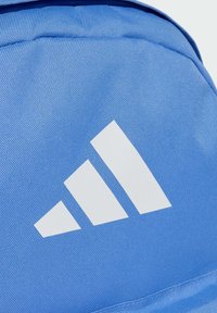 Mochila de tela azul con un prominente logo de tres franjas blancas. El material tiene una apariencia texturizada y duradera.