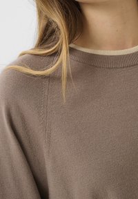 Gros plan d'une personne portant un pull tricoté taupe avec un col rond côtelé et des cheveux châtain clair reposant sur l'épaule.