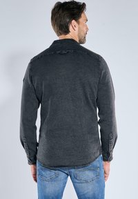 Ein Mann trägt ein dunkelgraues Hemd mit Knöpfen und blaue Jeans, steht mit dem Rücken zu einer schlichten hellgrauen Wand.