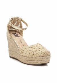 Sandalia de cuña beige con textura trenzada y un intrincado bordado. Cuenta con una correa al tobillo y una suela de tejido con apariencia desgastada.