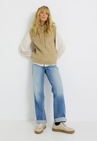 Haut sans manches en tricot beige avec des cordons de serrage sur une chemise à manches longues crème, associé à un jean droit bleu clair et des baskets beiges.