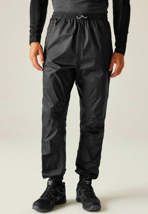 Mężczyzna ubrany w czarne nieprzemakalne spodnie i czarne buty turystyczne, stojący na tle jednolitego jasnego tła.