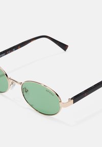 Green-tinted oval sunglasses with a gold metal frame, featuring black tortoiseshell-patterned arms and a logo on the lens.