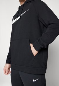 Sweat à capuche noir en tissu doux, avec un grand logo Nike Swoosh blanc sur la poitrine, une capuche réglable avec cordon de serrage, et une poche avant.