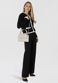 Cardigan nero con bordi bianchi, bottoni dorati; borsa crema con motivo logo e tracolla in argento; modella che indossa pantaloni neri e tacchi.