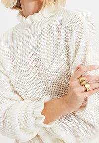 Bílý pletený svetr s volným vysokým límcem, dlouhými rukávy a texturovanou tkaninou. Má volný střih a žebrované manžety. Zlacené prsteny na ruce.