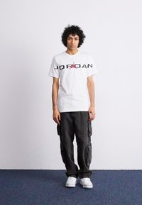 Tricou alb cu logo-ul "JORDAN" în culori negru și roșu, asortat cu pantaloni cargo negri și adidași albi. Coafat cu păr creț.