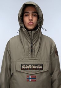 Beige gewatteerde anorak met een ritsnek, grote opgestikte zak en logoprint. Beschikt over een Noors vlagembleem, zwarte accenten en trekkoorden.