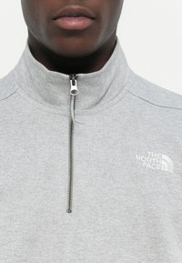 Felpa in pile grigia con colletto alto, chiusura con zip a un quarto e logo bianco sul lato sinistro del petto. Tessuto liscio e design casual.