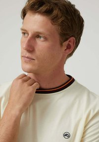 Camiseta de manga corta color crema con un cuello de canalé oscuro en contraste que presenta rayas rojas y amarillas. Logo circular en el lado izquierdo del pecho.