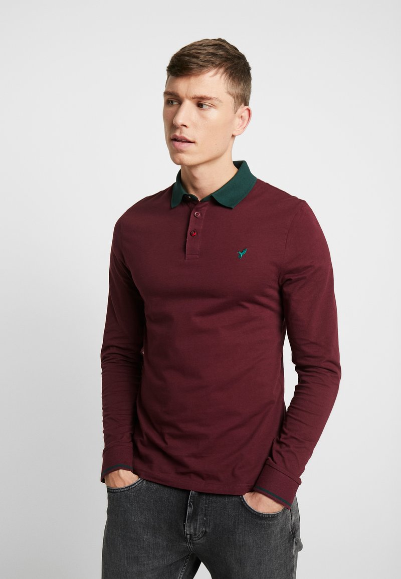 Polo à manches longues bordeaux avec un col vert, comportant deux boutons et un petit logo brodé sur la poitrine. Tissu doux.