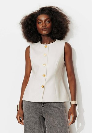 Femme aux cheveux bouclés et volumineux porte un haut blanc sans manches avec de gros boutons dorés, un jean gris, un collier en or et un bracelet assorti.