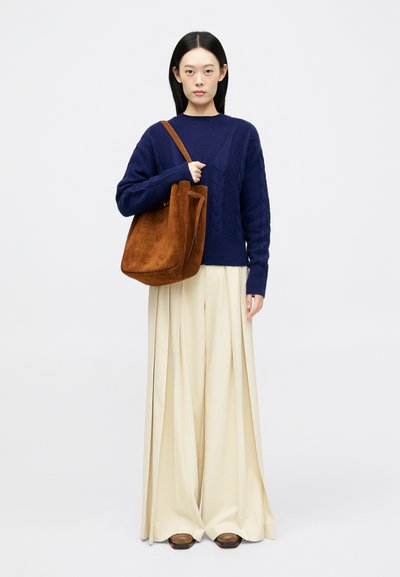 Ναυτικό πουλόβερ με πλέξη κασμίρι, κρέμα ψηλές παντελόνες και καφέ σουέτ τσάντα. Το μοντέλο στέκεται στο κέντρο, με τα χέρια πάνω στη θηλιές της τσάντας.