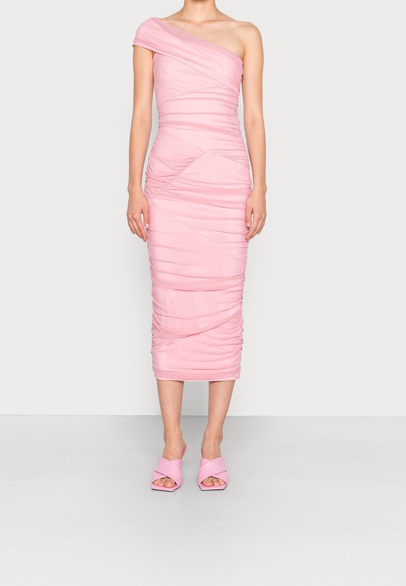 Femme debout portant une robe midi rose ajustée, froncée, asymétrique à une épaule, assortie à des mules à talons ouvertes rose, sur un fond uni.