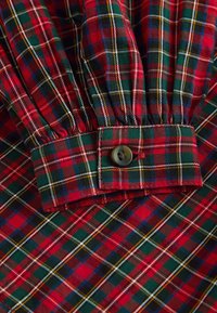 Tissu à carreaux rouge et vert avec des accents blancs et jaunes ; présente un poignet boutonné, une texture douce et un design froncé.