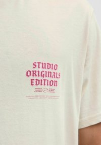 Krēms krāsas kokvilnas T-krekls ar drosmīgu rožainu izšūtu tekstu "STUDIO ORIGINALS EDITION" un papildus mazāku tekstu zem tā. Īsas piedurknes, parasts piegriezums.