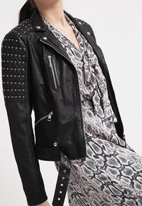 Giacca da moto in pelle nera con spalle borchiate, diverse cerniere e un design aderente. Indossata sopra un vestito con motivo a serpente.