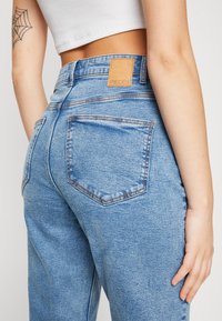 Jasnoniebieskie dżinsy z wysokim stanem i prostą nogawką. Posiadają tylne kieszenie i brązową skórzaną metkę z napisem "PIECES".