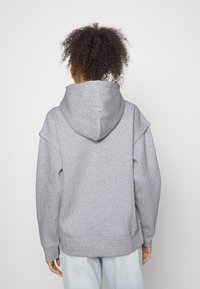 Sudadera gris con capucha y ajuste relajado, que presenta una capucha grande y diseño de hombros caídos. La tela tiene una textura suave y una construcción sencilla.