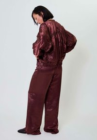 Vyno spalvos satino švarkas su didelėmis rankovėmis ir raukšlėtu nugaros uždengimu; atitinkamos plačios satino kelnės; lygus paviršius.