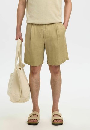 Personne portant un short beige et des sandales, tenant un sac fourre-tout couleur crème dans une main, l'autre main dans la poche.