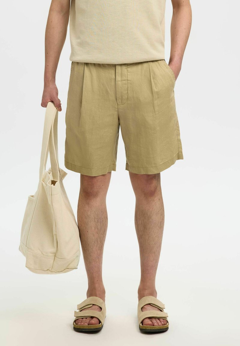 Personne portant un short beige et des sandales, tenant un sac fourre-tout couleur crème dans une main, l'autre main dans la poche.