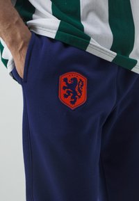 Námořnické tepláky s hladkou texturou, na kterých se nachází oranžový štítový logo se lvem a textem "KNVB". Viditelná boční kapsa na kyčli.
