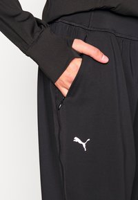 Svarta sportbyxor i lättviktig tyg, med en ficka med dragkedja, elastisk midja och en vit Puma-logga på sidan.