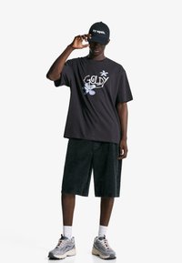 Schwarzes, übergroßes T-Shirt mit "GOLDY" und floralem Design, kombiniert mit dunkelblauen Denim-Shorts und grauen Sneakers. Schwarze Cap mit weißem Text.