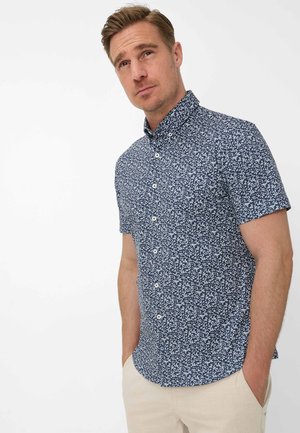 Mann trägt ein marineblaues kurzärmeliges Hemd mit weißen Blattmustern und hellbeige Hose, Hände in den Taschen.