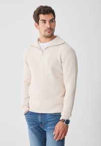 Ráncos krémszínű pulóver félzipz�árral és strukturált mintával, kék farmernadrággal és órával párosítva. Simma, puha anyag.