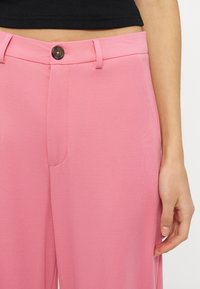 Κοντινή λήψη ενός ατόμου που φοράει ροζ παντελόνι με ψηλή μέση με μαύρο κουμπί και μαύρο κοντό τοπ, με το χέρι να ακουμπάει στο πλάι.