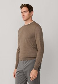 Bruine gebreide trui met een ronde hals en ribbel manchetten, gecombineerd met grijze broek. Subtiele branding op de borst. Zachte textuur.