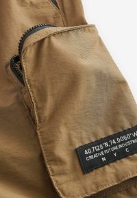 Brauner Stoff mit glatter Textur, der eine mit Reißverschluss verschlossene Tasche und ein schwarzes Etikett mit Koordinaten und Markennamen aufweist.
