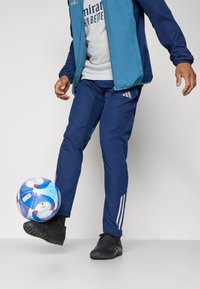 Blå adidas træningsdragt med hvide striber, der inkluderer en lynlåsjakke og tætsiddende bukser. Sorte fodboldstøvler og en mønstrede fodbold er til stede.