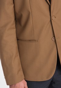 Ruskea bleiseri, jossa on jämäkkä muotoilu, kahdella napilla ja tyylikkäällä taskulla, jossa on kontrastivärinen koristelu, yhdistettynä tummiin housuihin.