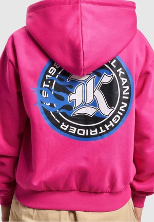 Sweat à capuche rose vif avec un grand logo circulaire bleu, noir et blanc présentant une lettre K et un motif de flammes au dos.