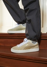 Baskets en daim beige avec des lacets blancs et un logo doré, posées sur un escalier en bois ; accompagnées de pantalons gris à bord élastique.