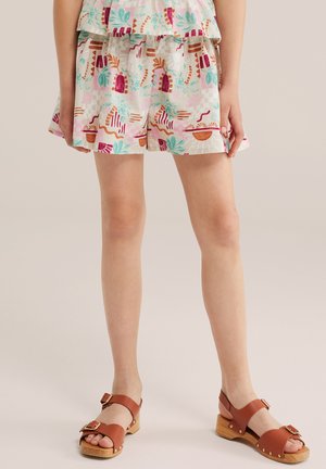 Enfant portant un short imprimé coloré et un haut assorti avec des sandales en bois marron à brides, debout devant un fond uni.