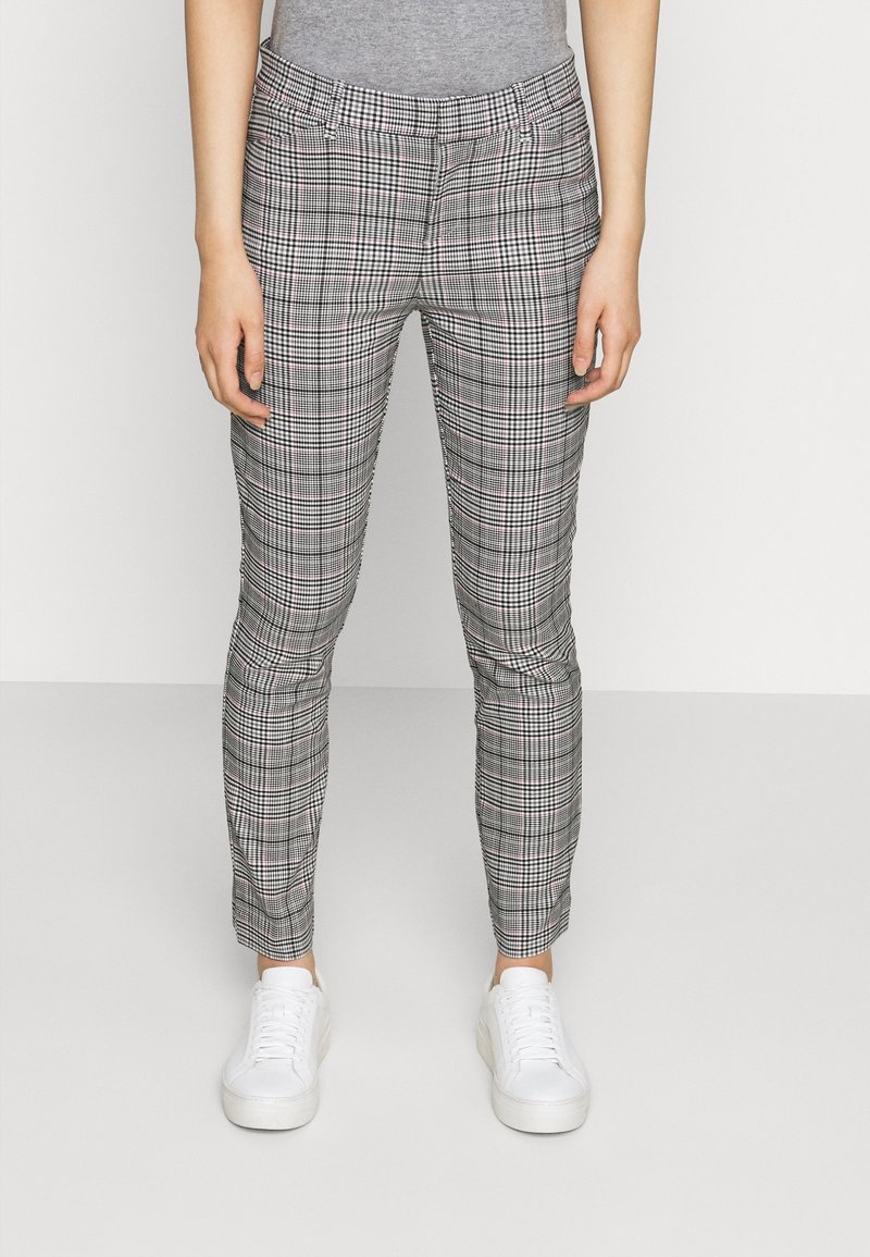 Personne portant un pantalon à carreaux gris ajusté à la cheville avec des baskets blanches, debout devant un fond uni clair.