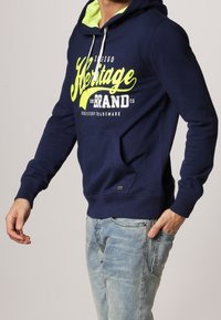 Mann in navy-blauem Hoodie mit neongelbem "Heritage Brand"-Text und hellblauen Jeans, Hand in der Tasche vor einfarbigem Hintergrund.