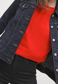 Giacca di jeans blu scuro con cuciture arancioni, abbottonata davanti e con due tasche sul petto, abbinata a un top rosso uniforme e pantaloni neri.